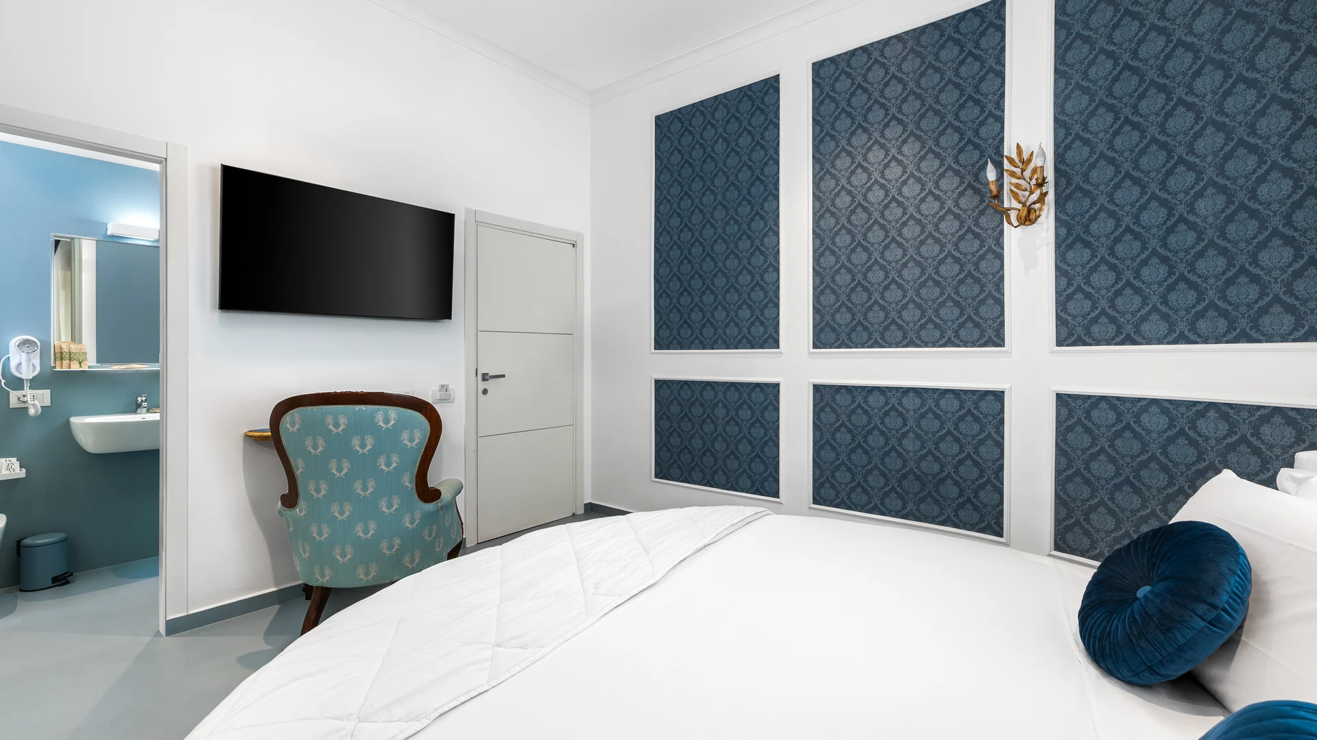 Camera d'hotel con letto bianco, poltrona azzurra e bagno con pareti blu