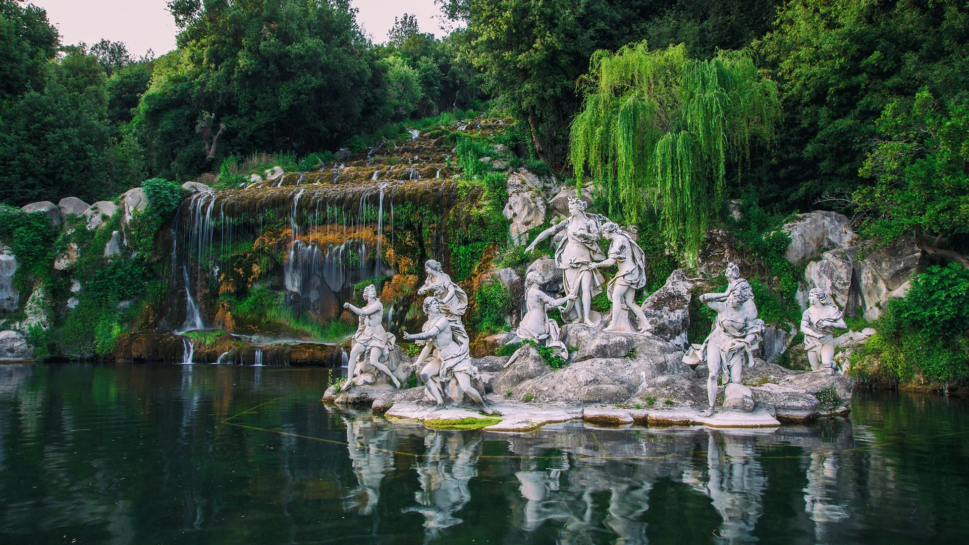 Statue di figure mitologiche su un'isola in un lago con cascata e vegetazione verde