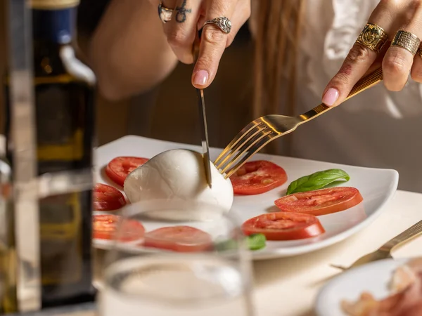Hotel Regina Caserta · Sapori di Campania Persona che taglia mozzarella circondata da fette di pomodoro e basilico su piatto bianco