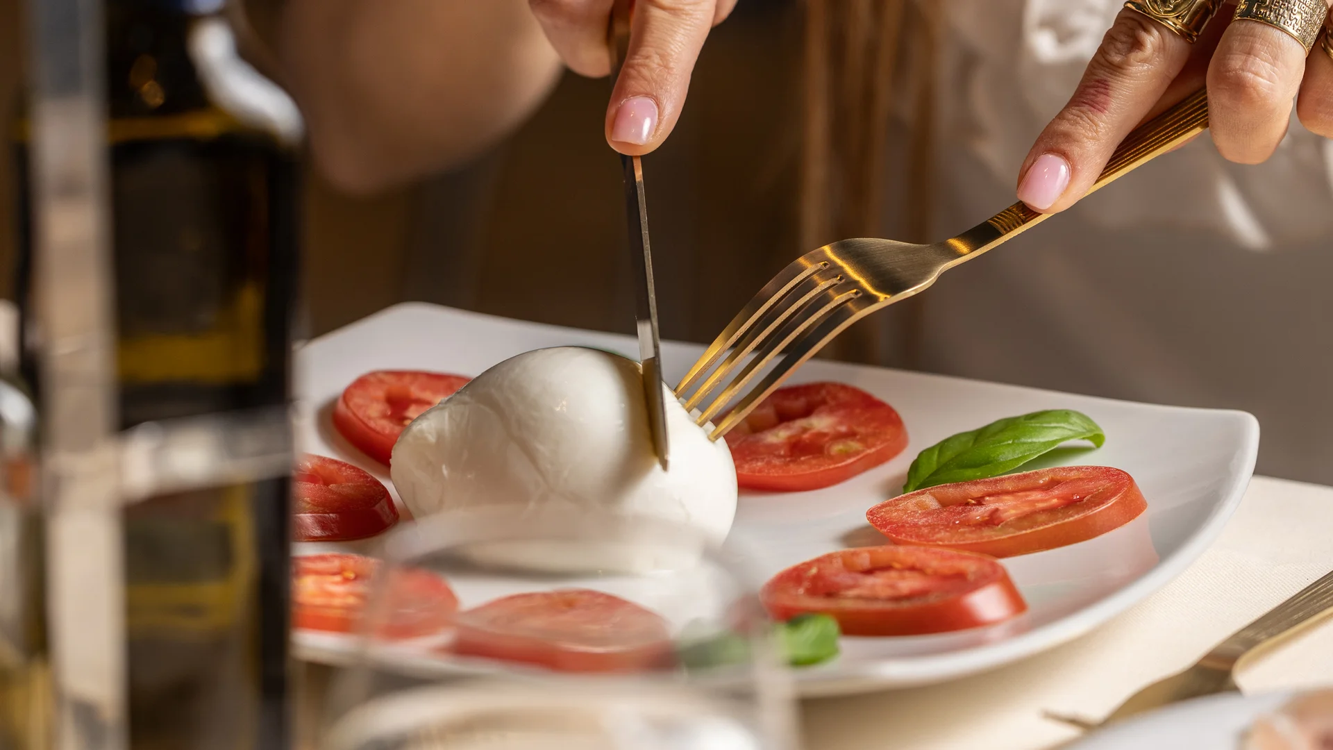 Persona che taglia mozzarella circondata da fette di pomodoro e basilico su piatto bianco