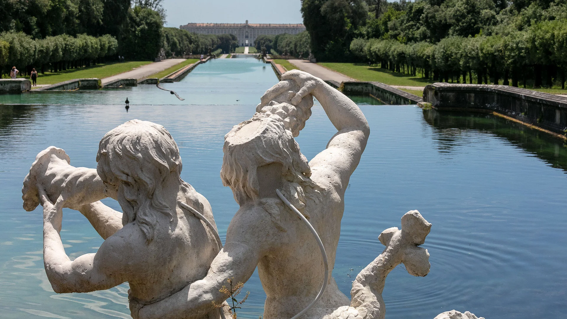 Statua di due figure mitologiche vicino a un lungo canale d'acqua con giardini e palazzo sullo sfondo