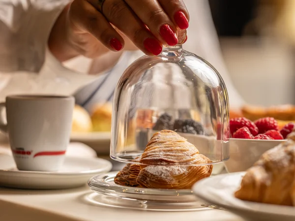 Mano con unghie rosse che solleva una campana di vetro su un croissant
