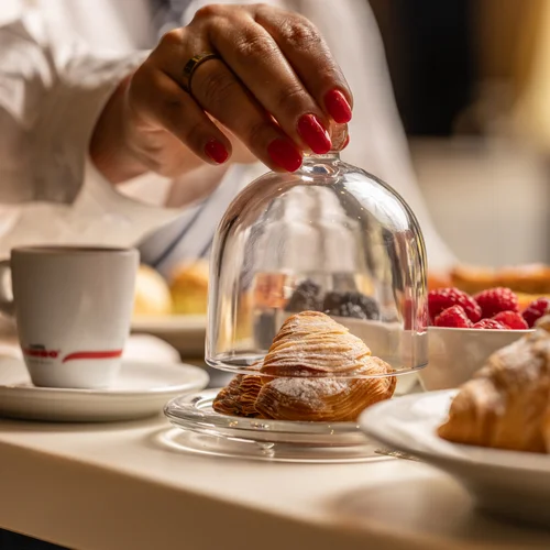 Mano con unghie rosse che solleva una campana di vetro su un croissant