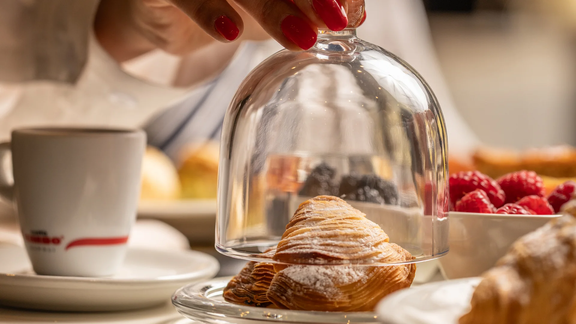 Mano con unghie rosse che solleva una campana di vetro su un croissant