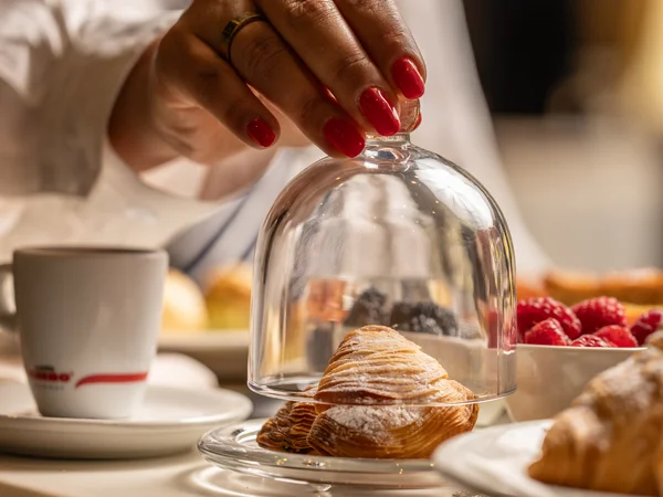 Hotel Regina Caserta · Sapori di Campania Mano con unghie rosse che solleva una campana di vetro su un croissant