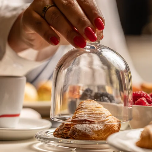 Hotel Regina Caserta · Sapori di Campania Mano con unghie rosse che solleva una campana di vetro su un croissant