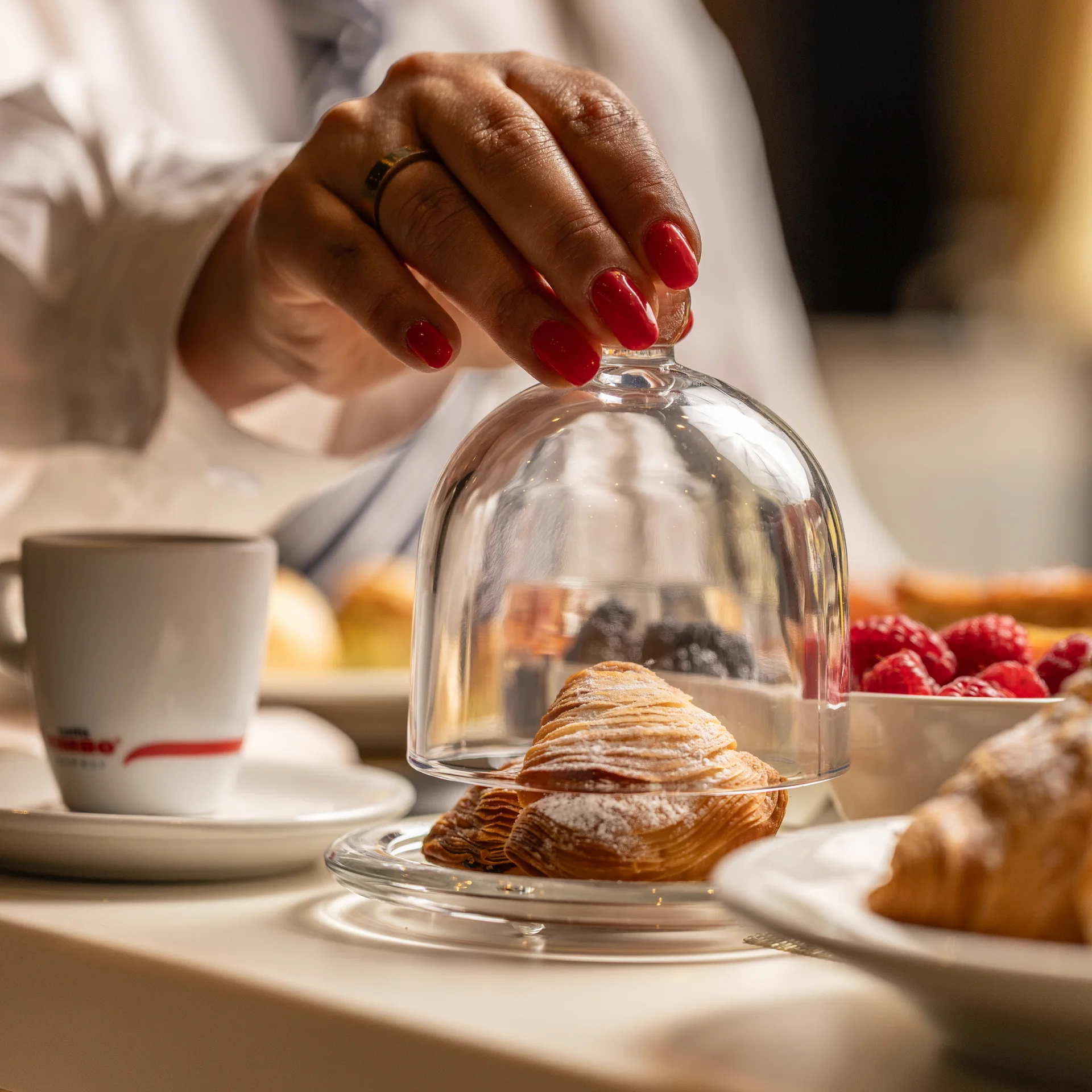B&b a Caserta vicino alla Reggia · La colazione reale Mano con unghie rosse che solleva una campana di vetro su un croissant