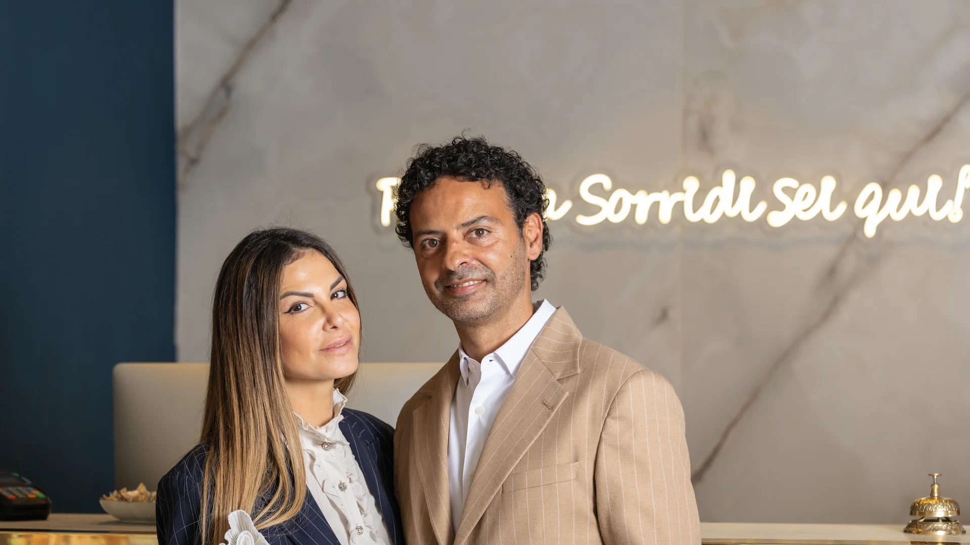 Hotel Regina Caserta · La nostra filosofia di ospitalità Coppia elegante sorridente alla reception di un hotel con scritta luminosa sullo sfondo